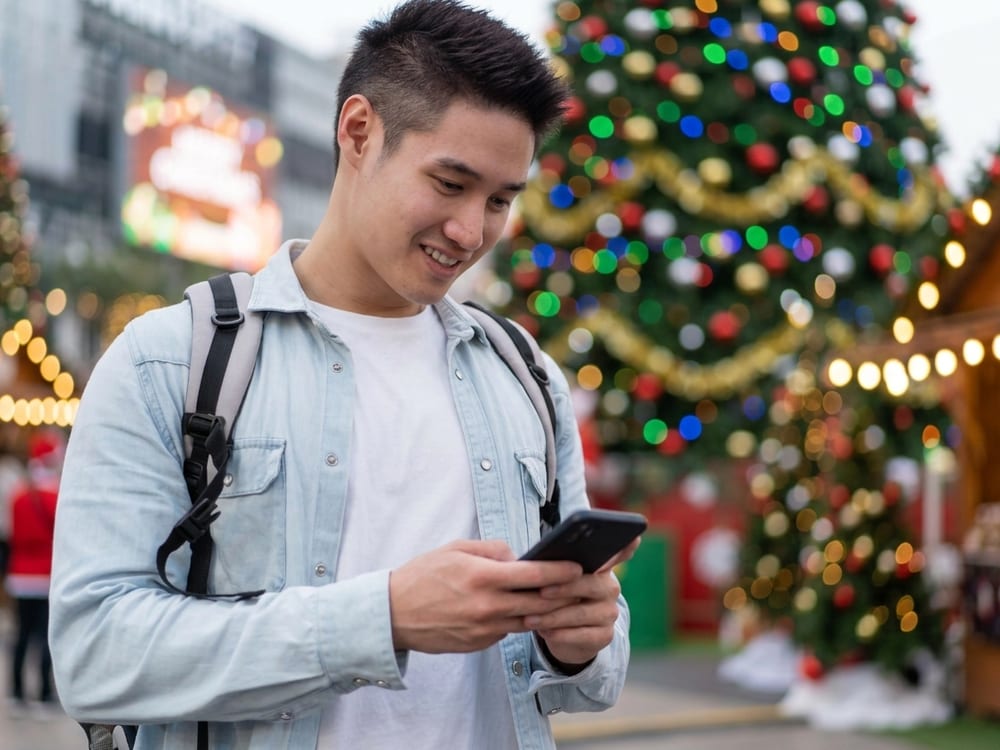 A young man at a Christmas market beside a lit tree, smiling at something on his phone